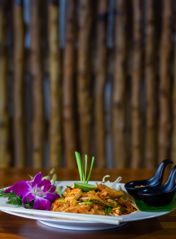 about-us-02 Close-up of a colorful Thai Pad Thai dish garnished with an orchid, captured indoors.